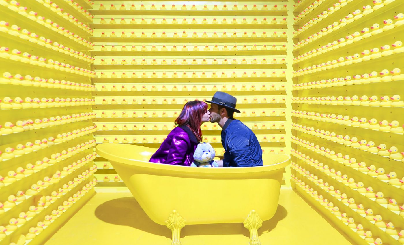 Para ubrana na niebiesko i fioletowo siedzi w żółtej wannie i otaczające ściany również są żółte
