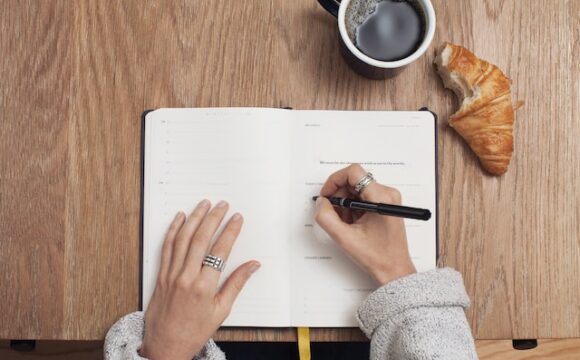 Dłonie kobiety piszącej dziennik. Obok stoi kubek kawy.