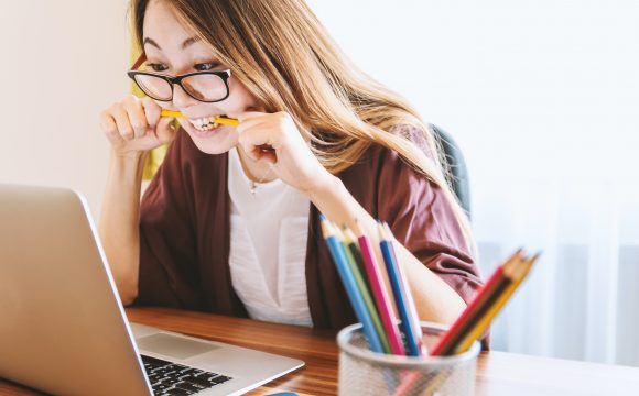 Kobieta w okularach gryzie ołówek siedząc przed otwartym laptopem
