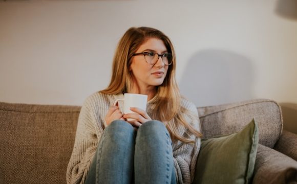 Kobieta w okularach trzymająca w dłoniach białą filiżankę, siedzi na sofie z podkulonymi nogami