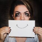 Kobieta z rozmazanym tuszem wokół oczu trzyma kartkę z narysowanym uśmiechem