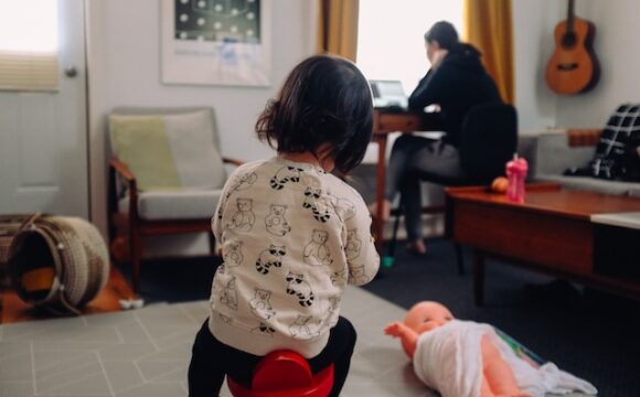 Dziecko w białej bluzce siedzi na krzesełku a przy biurku siedzi mama pracująca zdalnie.