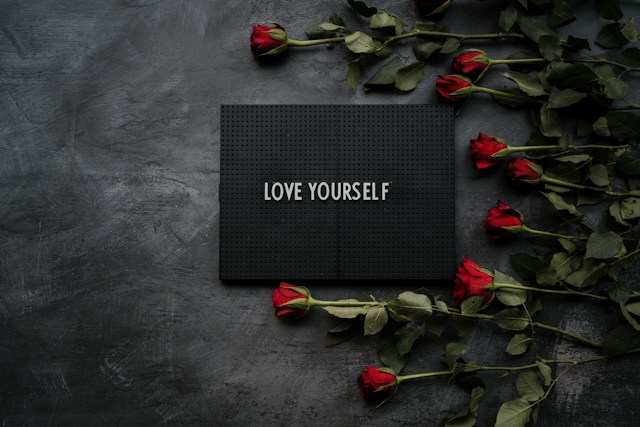 Czerwone róże na ciemnym tle, a na środku czarna kartka z napisem kochaj siebie.