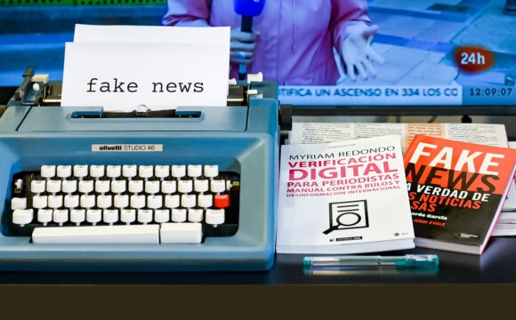 Maszyna do pisania, włączony telewizor oraz książki ostrzegające przed fake newsami