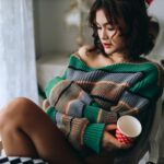 Kobieta owinięta w kolorowy koc i trzymająca kubek siedzi zamyślona na krześle