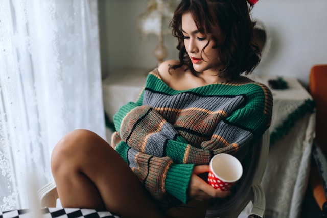 Kobieta owinięta w kolorowy koc i trzymająca kubek siedzi zamyślona na krześle