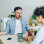 Mężczyzna w niebieskiej koszuli mówi do siedzącej obok kobiety, gestykulując