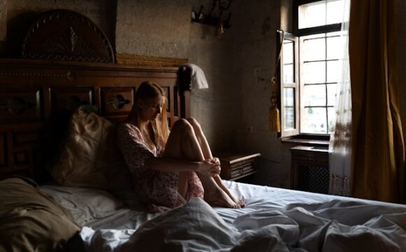 Kobieta w białej sukience siedząca na niezasłanym łóżku