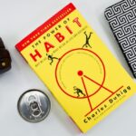 Żółta ksiażka o tytule The power of habits (siła nawyków) leży na stole obok słychawek i puszki