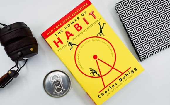 Żółta ksiażka o tytule The power of habits (siła nawyków) leży na stole obok słychawek i puszki