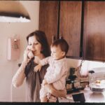 Kobieta w szarej bluzce trzyma dziecko na rękach i rozmawia przez telefon