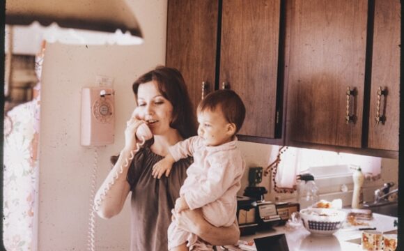 Kobieta w szarej bluzce trzyma dziecko na rękach i rozmawia przez telefon