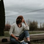 Kobieta w swetrze trzyma ksiażkę i siedzi zamyślona na poboczu drogi