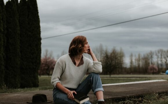 Kobieta w swetrze trzyma ksiażkę i siedzi zamyślona na poboczu drogi