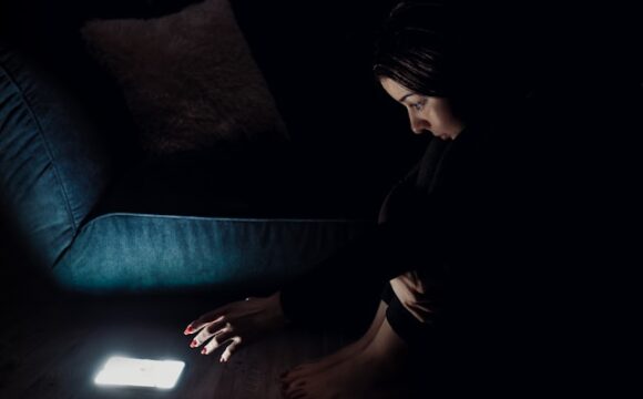 Kobieta siedząca na podłodze i wyciągająca ręce w stronę jasno świecącego smartfona