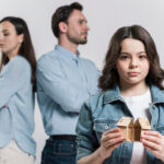 Smutna dziewczynka w jeansowej kurtce trzyma domek podzielony na pół, w tle stoją rodzice odwróceni do siebie plecami