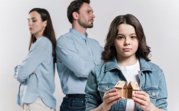 Smutna dziewczynka w jeansowej kurtce trzyma domek podzielony na pół, w tle stoją rodzice odwróceni do siebie plecami