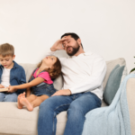 Zmęczony ojciec w białym swetrze siedzi na kanapie obok dwójki małych dzieci.