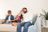 Zmęczony ojciec w białym swetrze siedzi na kanapie obok dwójki małych dzieci.