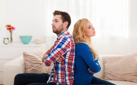 Kobieta w niebieskiej bluzce i mężczyzna w koszuli w kratkę siedzą z założonymi rękoma i są odwróceni do siebie plecami