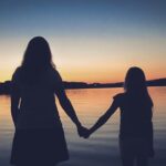 Sylwetki matki i córki trzymających się za ręce, na tle zachodzącego słońca.