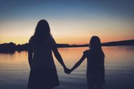 Sylwetki matki i córki trzymających się za ręce, na tle zachodzącego słońca.