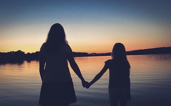 Sylwetki matki i córki trzymających się za ręce, na tle zachodzącego słońca.