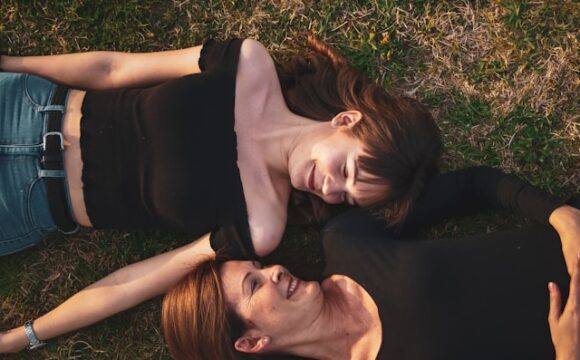 Dwie kobiety ubrane w czarne blizki leżą na trawie, patrzą i uśmiechają się do siebie