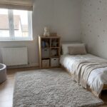 Umeblowany pokój utrzymany w tonacji bieli. Znajduje się w nim dywan, łóżko oraz mała szafka na rzeczy